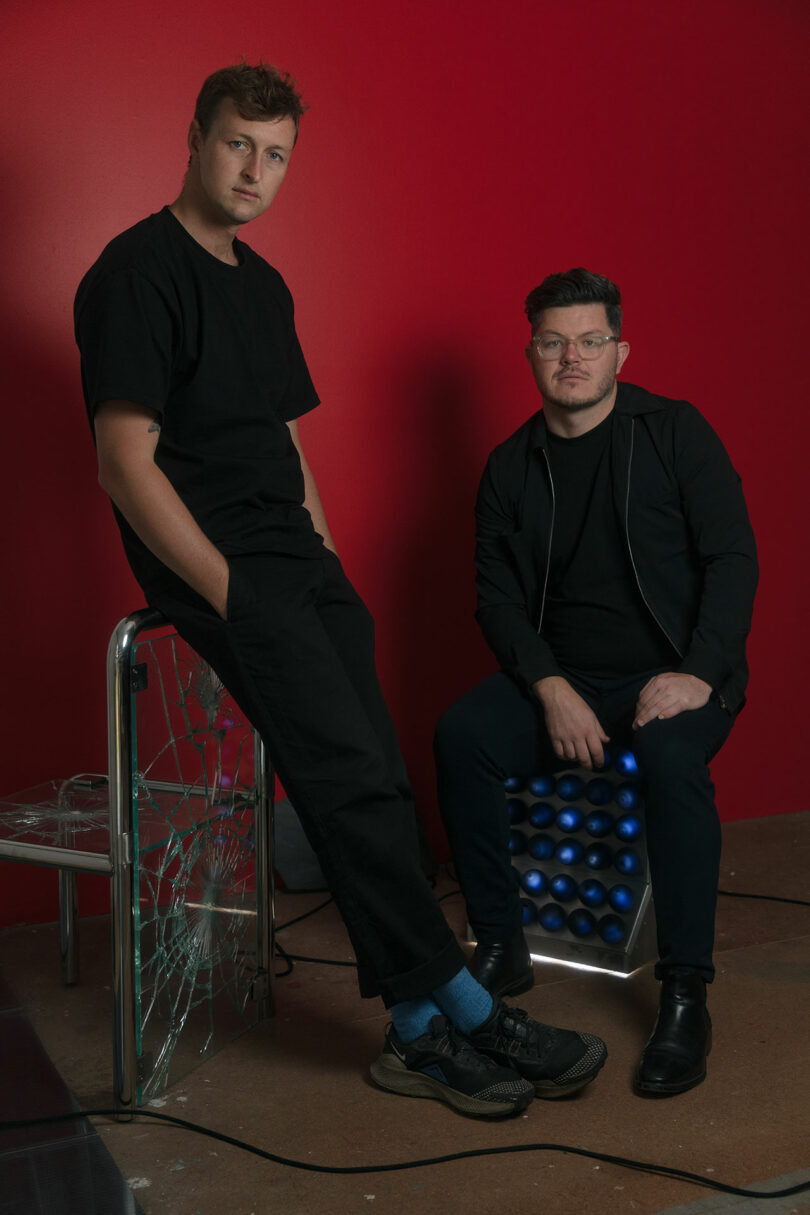 Two men in black clothing pose against a red wall; one, like a modern-day rider, sits on a glass chair while the other contemplates abstract objects for thought on a metal seat with blue lights.