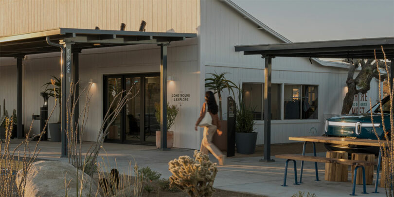 A person walks in front of a modern building with a light exterior, large windows, and plants. A covered outdoor area has benches and a table.