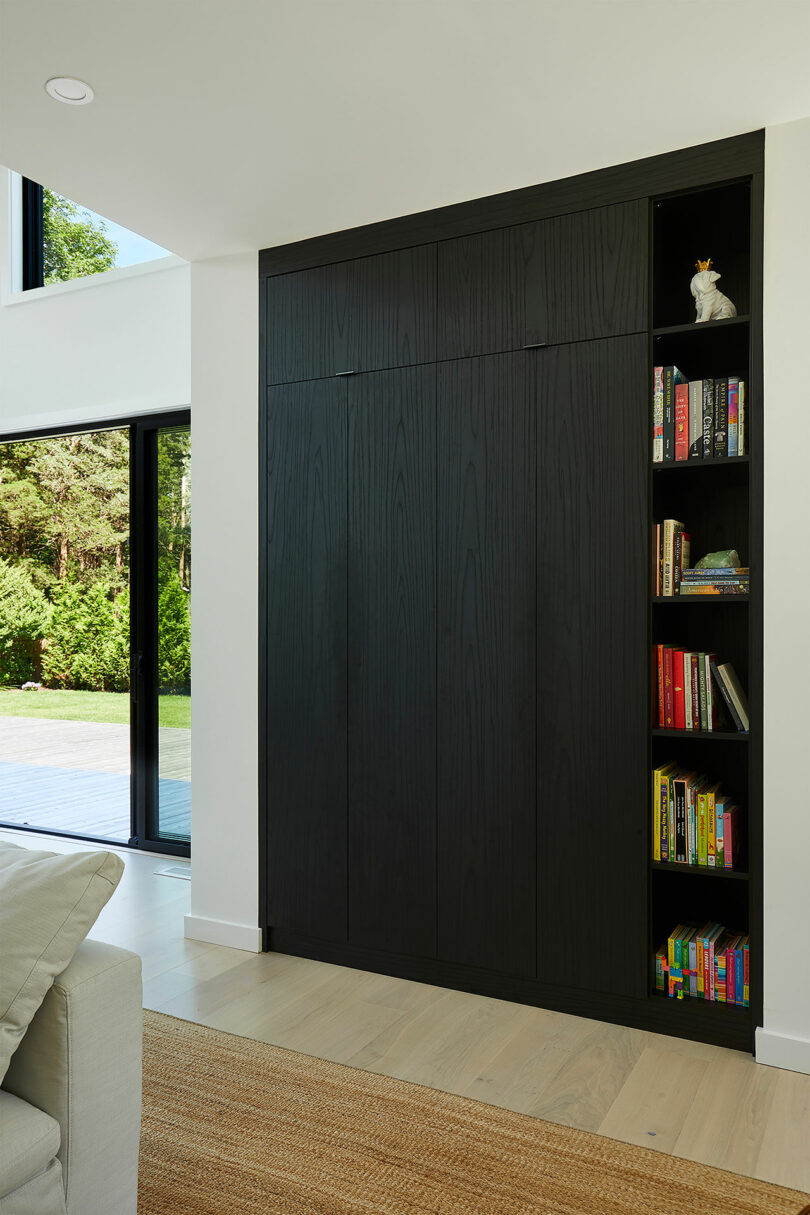 A modern living room in the Shift House features a large black cabinet with a built-in bookshelf filled with colorful books. Large windows reveal a stunning view of greenery outside, while a light gray couch is partially visible.