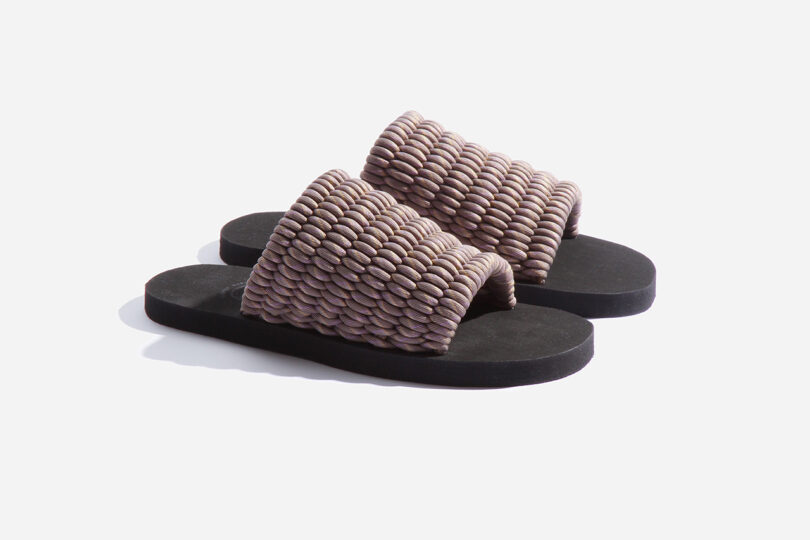 A pair of slide sandals with textured, woven upper straps in a light brown color and black soles, set against a plain white background.