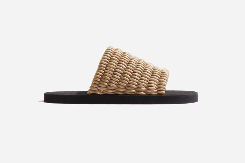 A single slide sandal with a wide, textured beige strap and a black flat sole, viewed from the side against a white background.