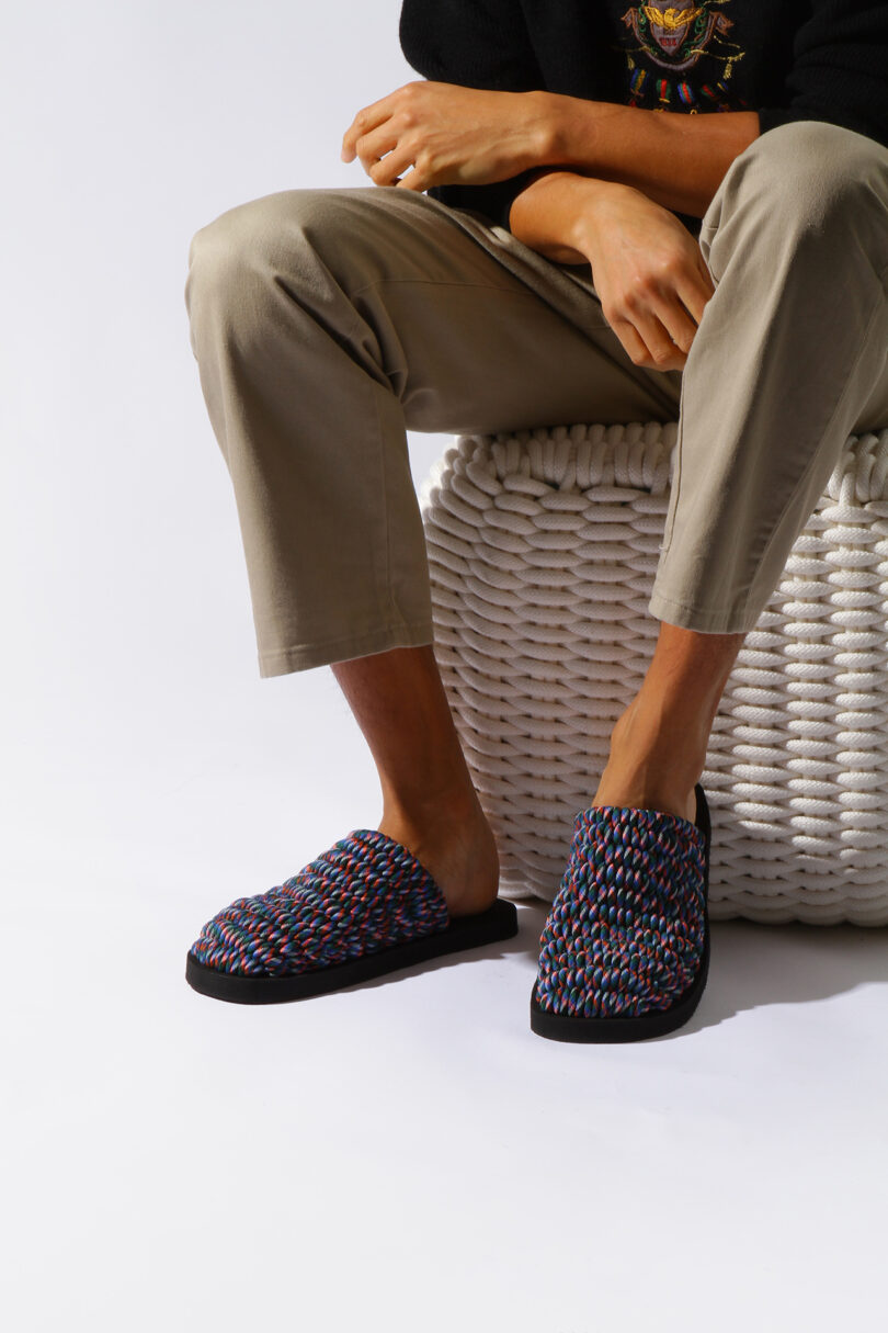 Person seated on a woven stool, wearing a dark sweater, beige pants, and colorful woven slippers, against a plain background.