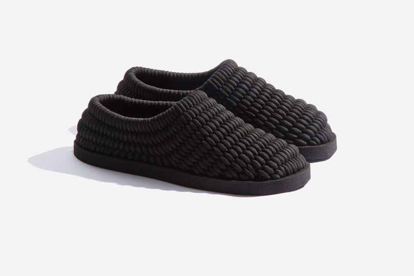 A pair of black slippers with a textured, braided design and flat soles, placed on a white background.