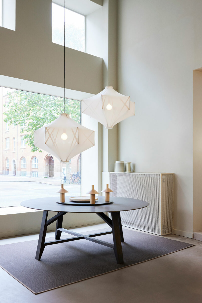 The modern dining room features a modern vibe with a round table, geometric pendant lights, and large windows. Neutral colors dominate the decor, including a textured rug and minimal accessories on the table.