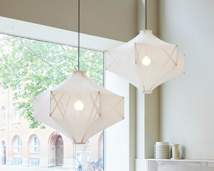 Two white geometric pendant lamps hang near a window with a view of brick buildings and vibrant greenery outside.