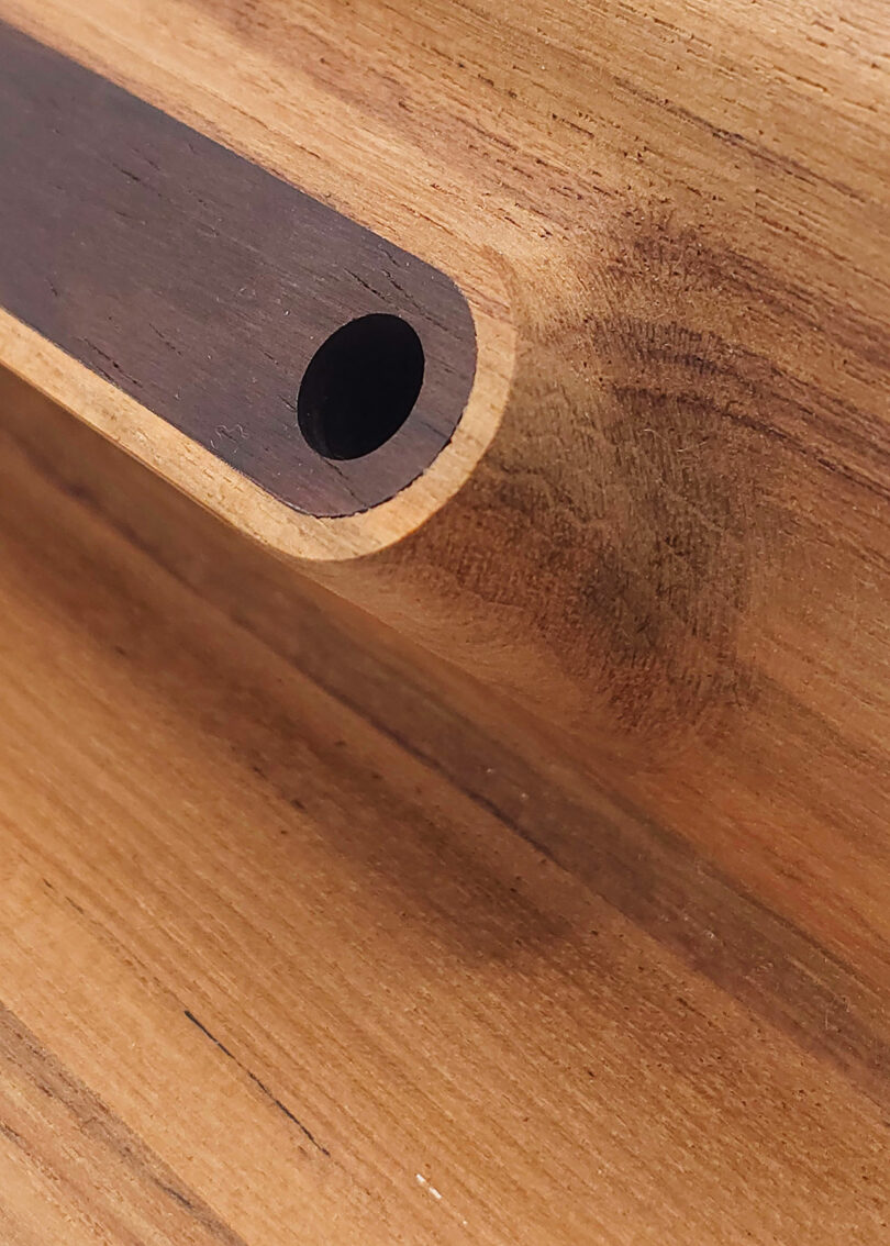 Close-up of a wooden surface with a rounded, dark groove containing a circular hole. The grain of the wood is visible, with tones of light and dark brown.