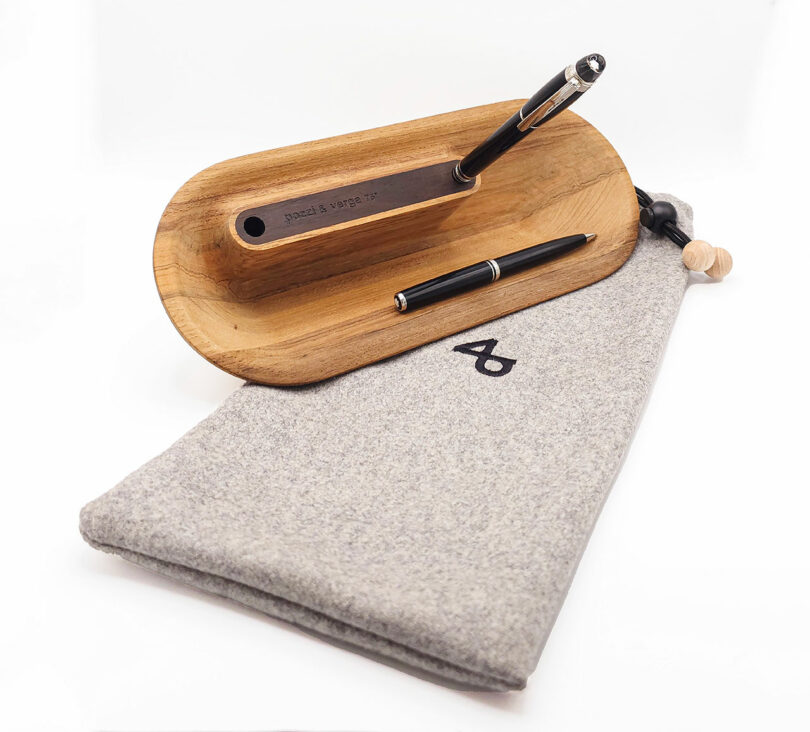 Wooden pen stand with two pens on it next to a gray fabric pouch.