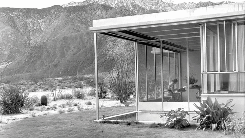 In a modern glass-walled house, a person pauses to take five amidst the sweeping desert landscape, with majestic mountains standing sentinel in the background.