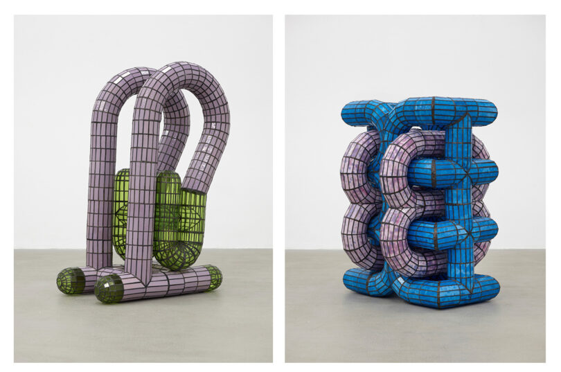 Aria Lee's Take 5 brings to life two abstract sculptures with interconnected tubular forms. The left sculpture is purple and green; the right, blue and purple. Both boast a grid pattern and stand on a plain floor, capturing an intriguing dance of colors and shapes.