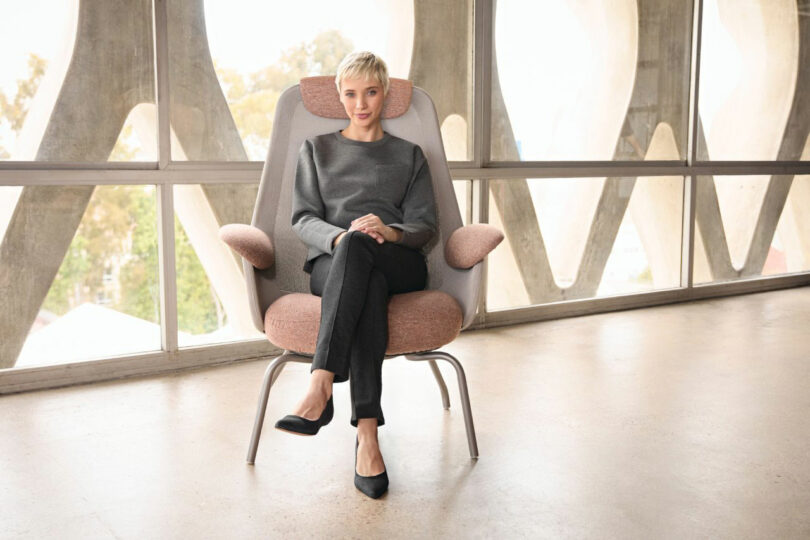 A person with short blond hair sits cross-legged on a sleek Teknion chair in a room with large windows.