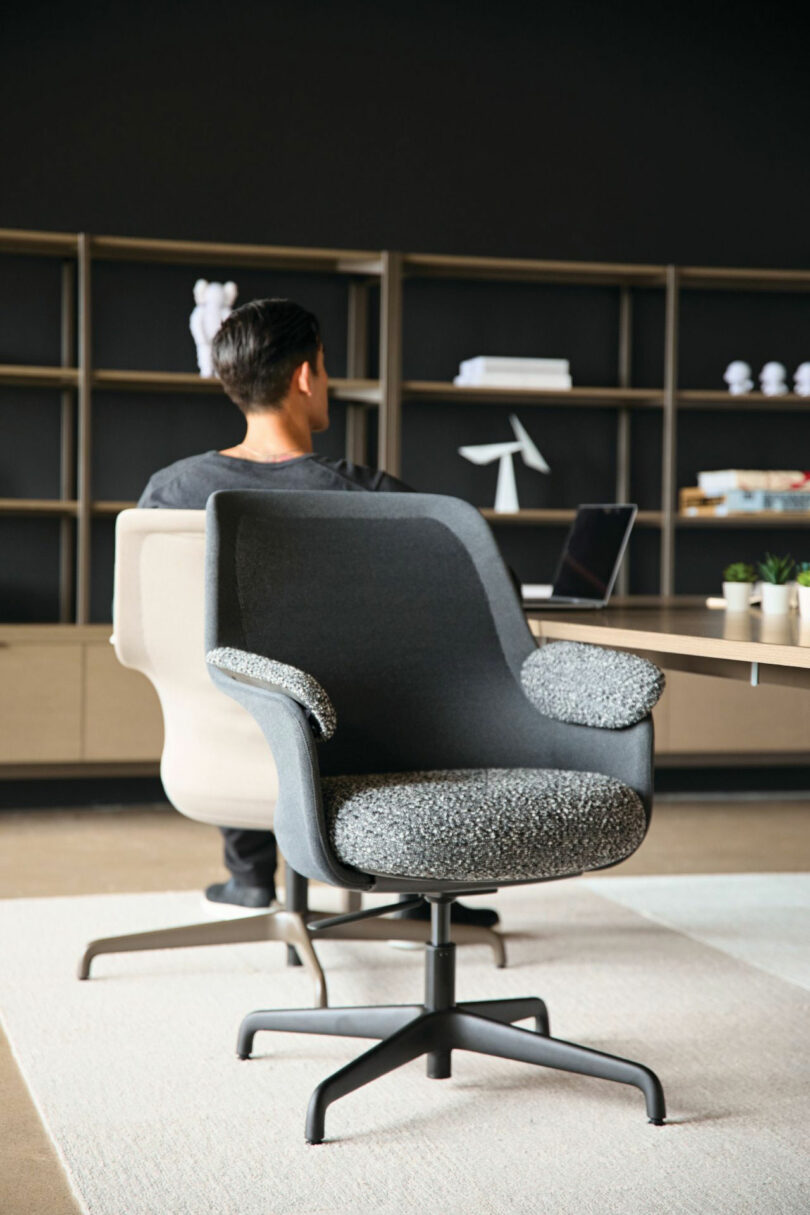 A man sits in a Teknion office chair at a desk with a laptop, facing away. Another empty chair is nearby. Shelves with books and plants create a serene backdrop, adding warmth to the workspace.