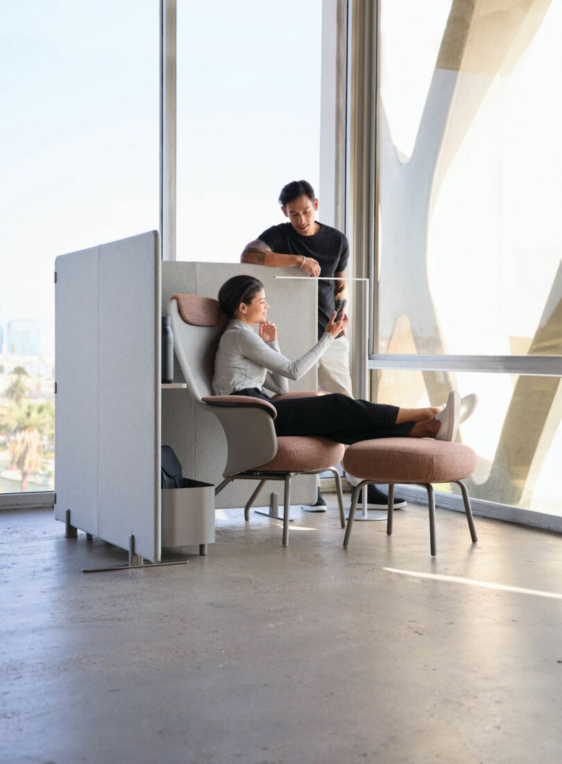 Two people in a sleek Teknion office space: one sits on a chair engrossed in their phone, while the other casually leans on a partition, observing. Behind them, floor-to-ceiling windows flood the room with natural light.