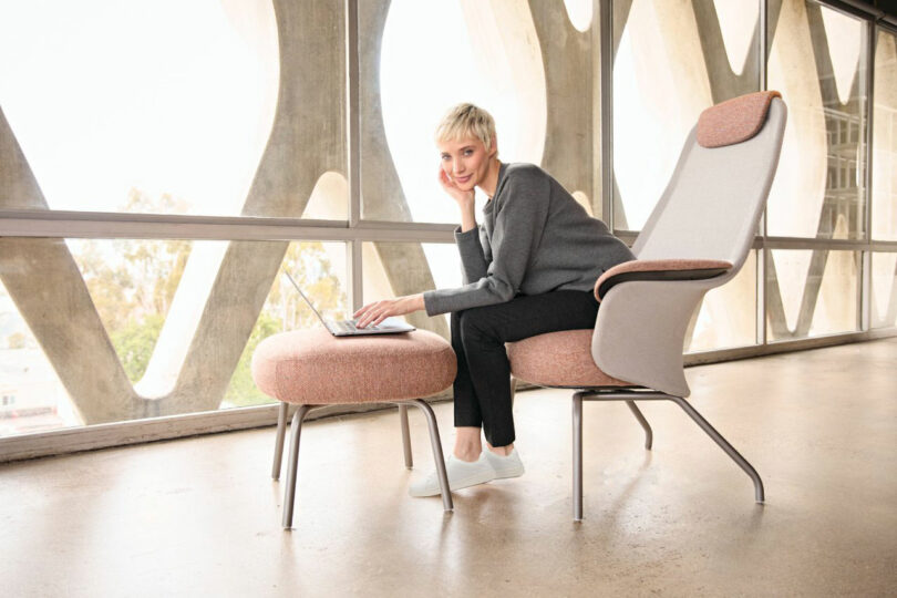 Seated in a sleek Teknion chair, the person uses a laptop resting on a footrest, basking in sunlight filtering through windows with unique geometric patterns.