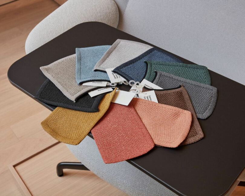 A variety of rectangular fabric swatches in different colors are fanned out on a black table next to a beige Teknion chair.