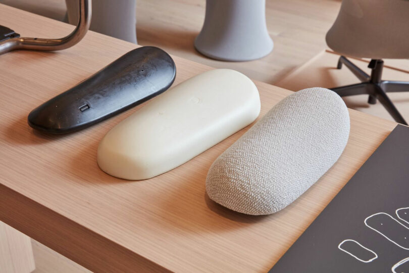 Three oblong objects are displayed on a sleek Teknion wooden table: one black, one white, and one textured gray.