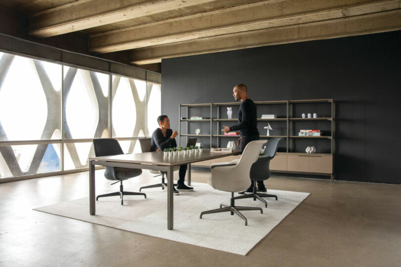 Two people converse in a modern Teknion office, featuring a large window and dark wall. They sit and stand around a table on a white rug, with shelves adorned with decorative items in the background.