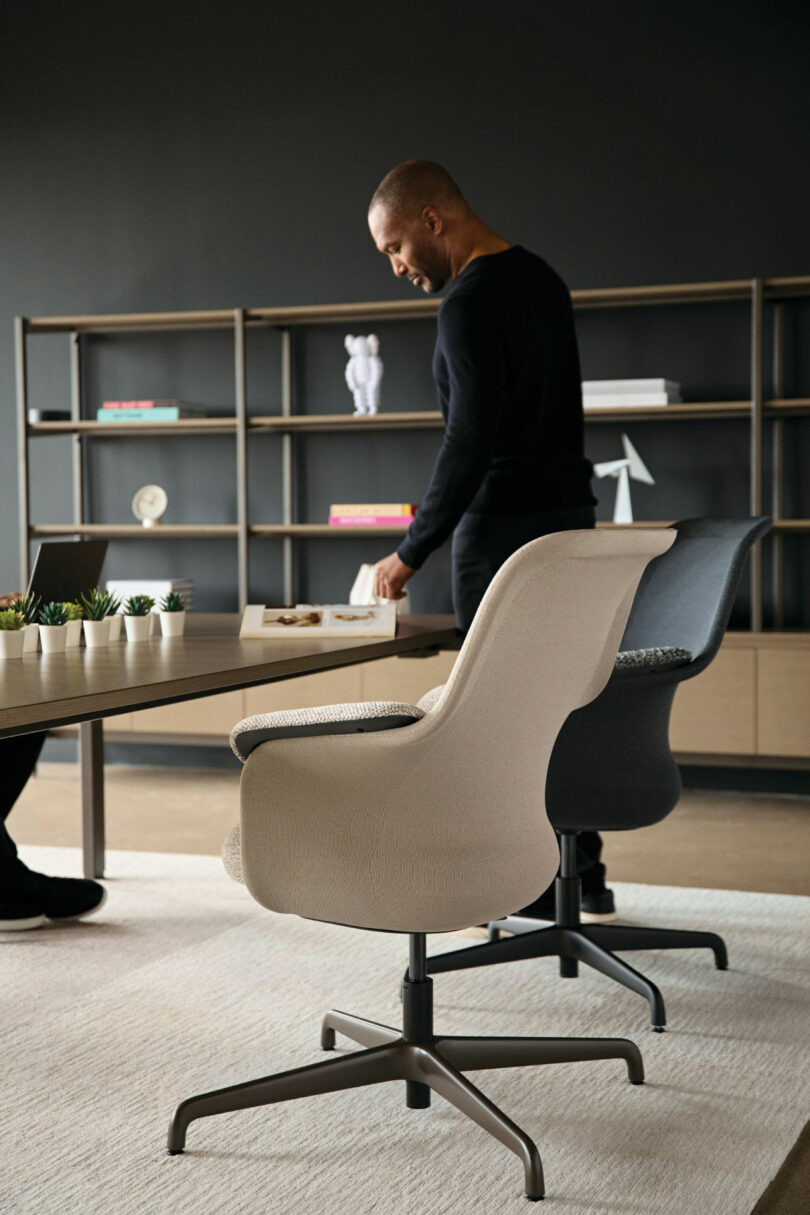 A person stands near a Teknion table in an office setting, with chairs and a bookshelf beautifully arranged in the background.