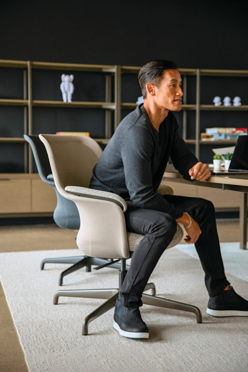 Seated in a modern office outfitted with sleek Teknion furniture, the person wears a dark sweater and pants, gazing attentively at a computer screen. Behind them, bookshelves and stylish office decor complete the contemporary ambiance.