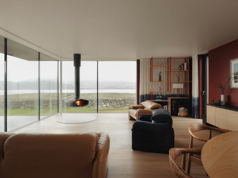 Modern living room with large glass walls showcasing an outdoor view, featuring a suspended fireplace, brown leather furniture, and a red accent wall with shelves.