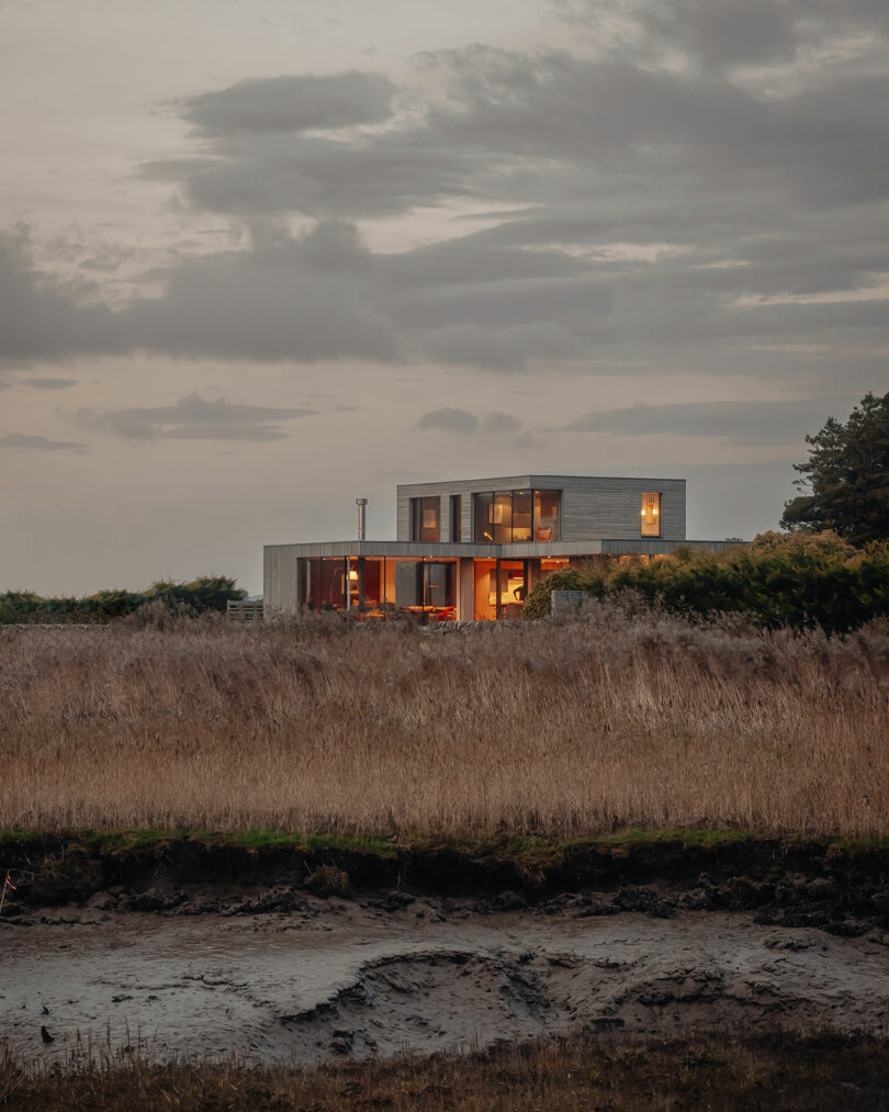 Modern two-story house with large windows, situated in a grassy field at dusk, with a cloudy sky above.