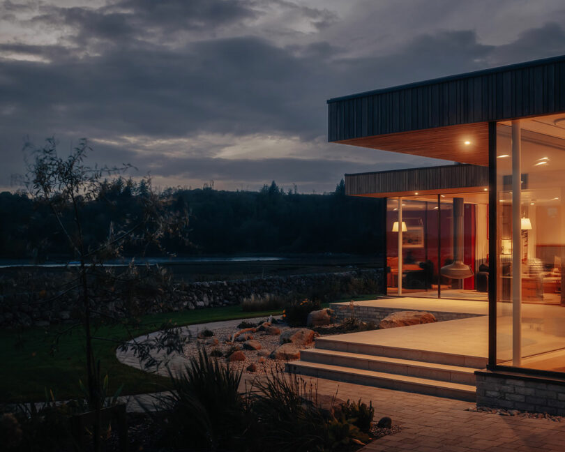 A modern house with large glass windows glows warmly at dusk. The exterior features a landscaped garden and stone pathway, with trees and hills visible in the background under a cloudy sky.