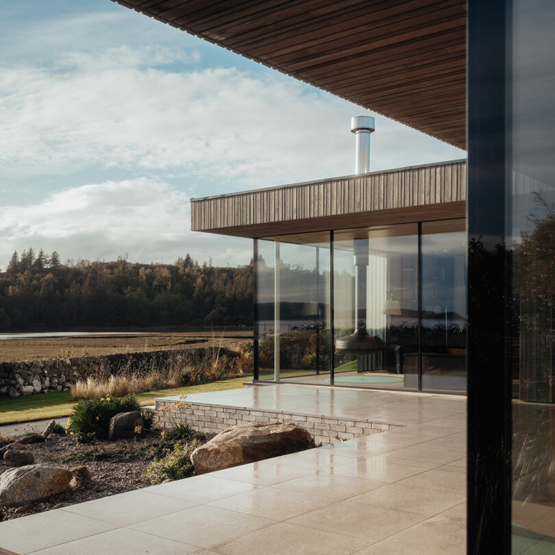 Modern house with large glass windows, sleek wood paneling, and a tiled patio. It overlooks a serene landscape with trees and a stone wall under a partly cloudy sky.