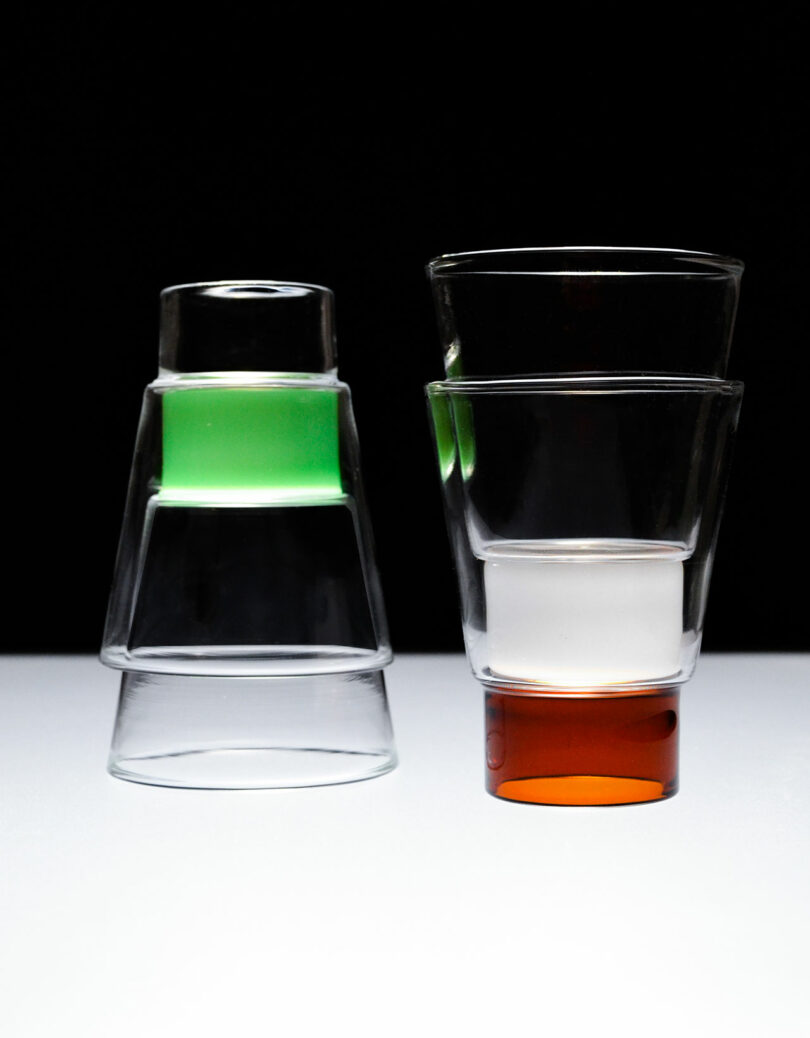 Two stacks of glasses on a white surface and black background. In a nod to the Podio style, one stack boasts a green liquid glass on top, while the other features a clear liquid in the middle and orange at the bottom.