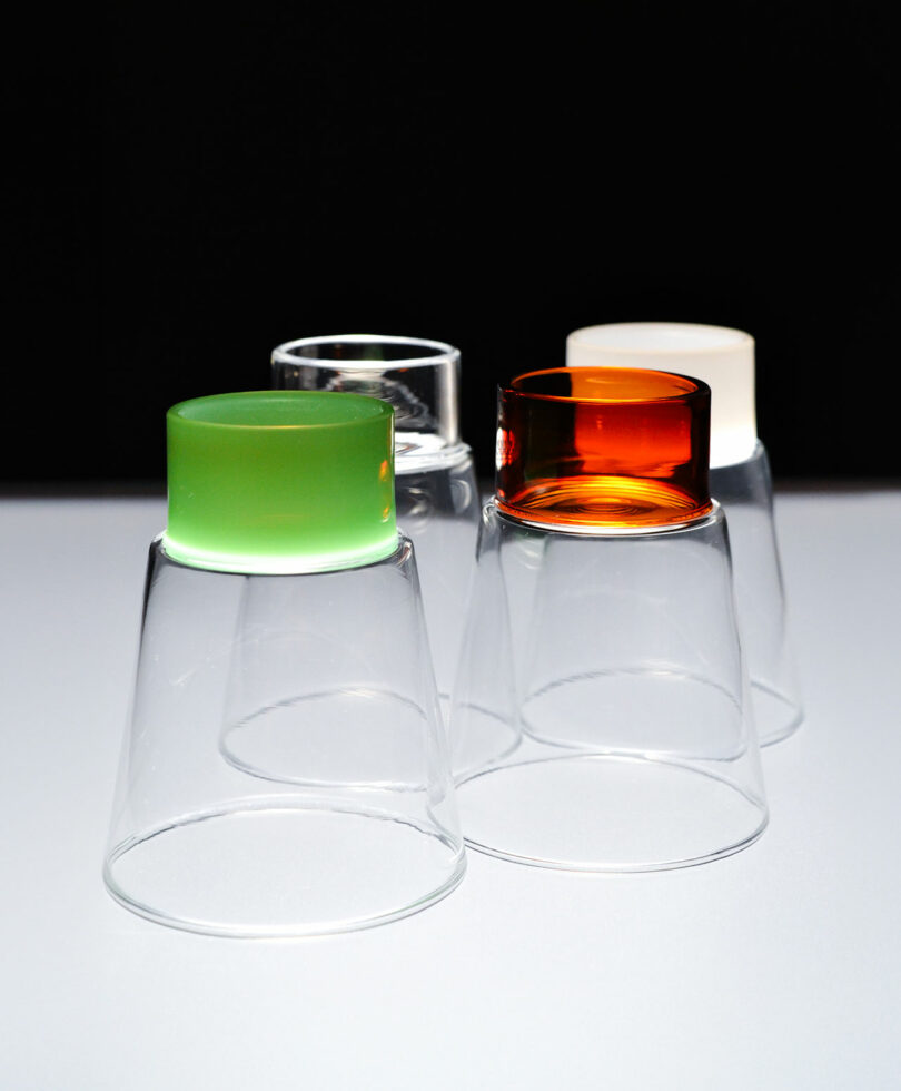 Four upside-down transparent glasses with vibrant Podio Cup bases in green, clear, orange, and white rest elegantly on a white surface against a stark black background.