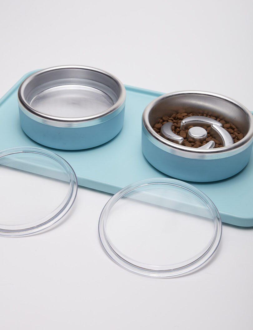 Two pet food bowls with lids on a blue mat; one bowl is empty, and the other contains dry pet food.