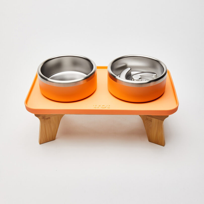 Orange elevated pet feeder with two stainless steel bowls. Left bowl is empty; right bowl has a spiraled slow feeder insert. Wooden stand elevates the feeder.