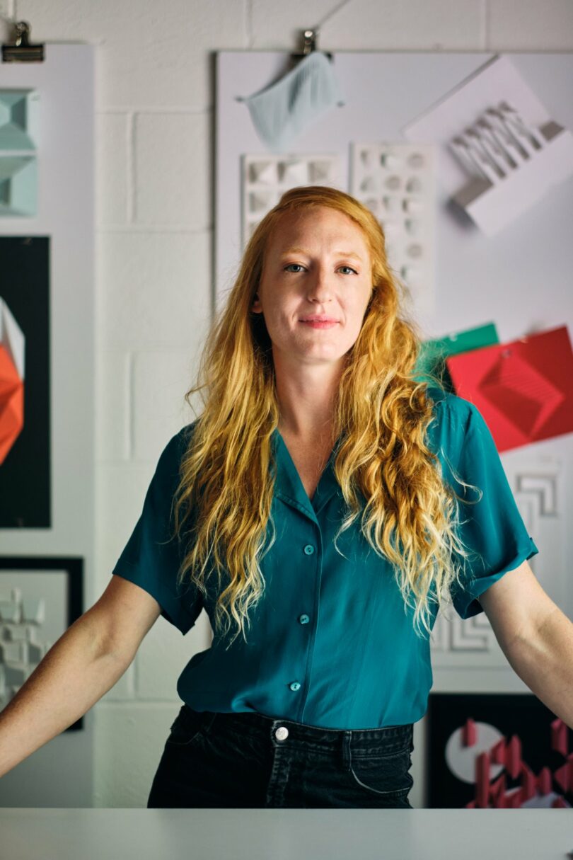 Person with long hair stands in front of geometric artworks on a wall.