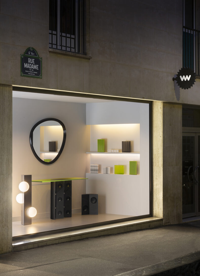 Minimalist store window display with a circular mirror, geometric sculptures, books, and glowing spherical lights on Rue Madame.