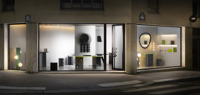 Storefront with large windows displaying modern furniture and decorative items, including geometric shapes and mirrors, under soft lighting at night.