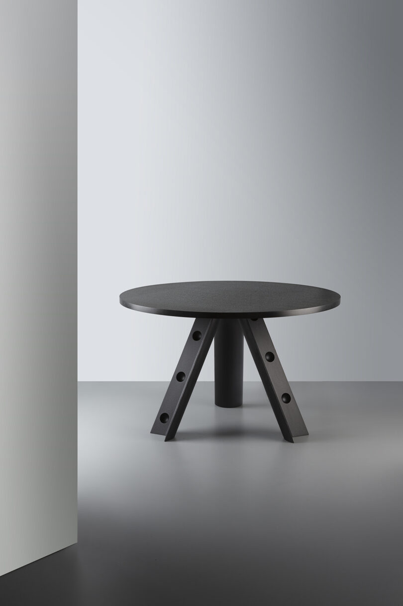 A modern round black table with a unique leg design is set against a minimalist background.