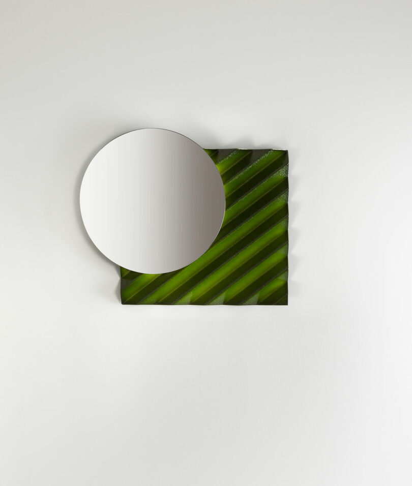 A concav design, inspired by Victoria Wilmotte, features a round mirror partially overlapping a green, diagonally striped square mounted on a plain white wall.