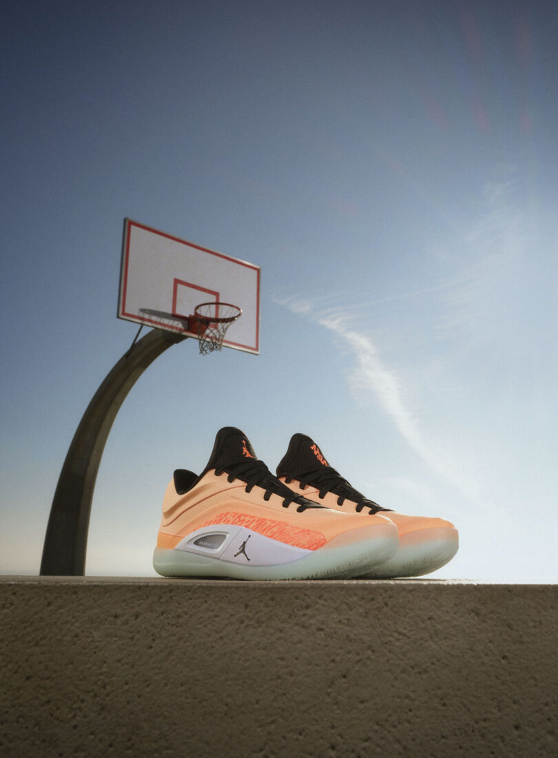 Une paire de baskets orange et noires repose sur une surface en béton avec un panier de basket en arrière-plan sur un ciel clair.