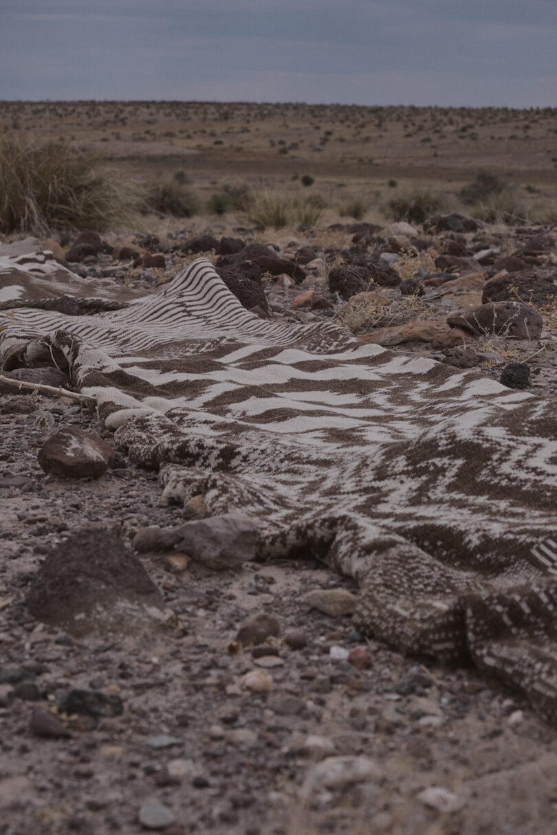 Patterned blanket lying on rocky desert ground, with distant shrubs and cloudy sky in the background