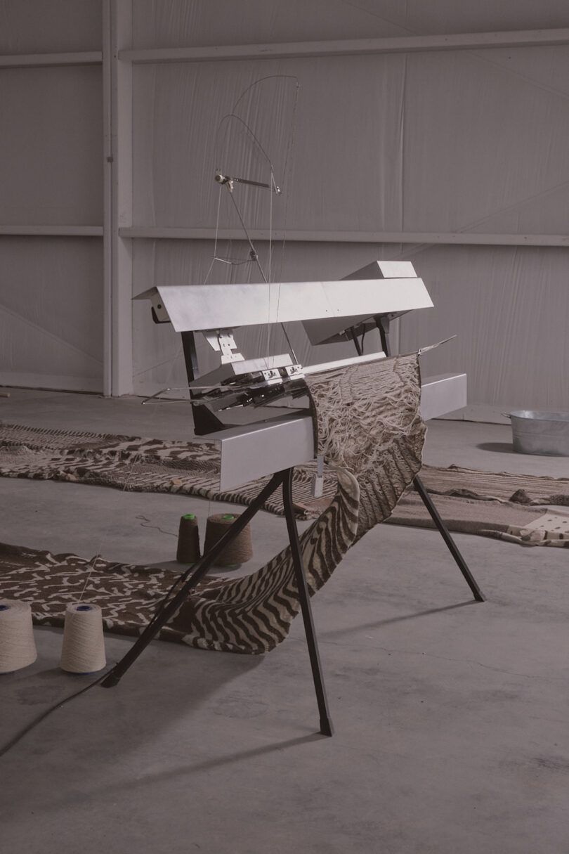 A modern art installation: a kniting machine on a tripod base with patterned fabric draped over it, situated in an industrial-style space with thread spools nearby