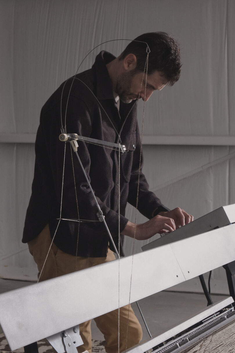 A man wearing a dark jacket and beige pants plays a unique stringed instrument in a dimly lit room