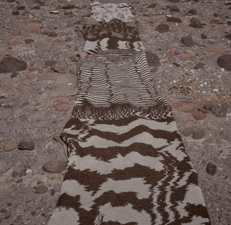 A long, patterned textile lies on rocky ground, featuring brown and beige wavy designs