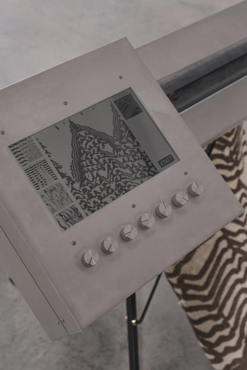 Close-up of a knitting machine interface displaying a patterned design on a screen, with six round knobs below. A partially knitted fabric in a similar pattern is visible