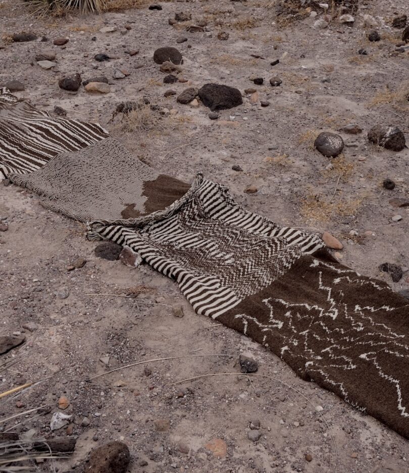 A long, textured fabric with various brown and cream patterns is spread across rocky, arid ground alongside some dry plants and stones