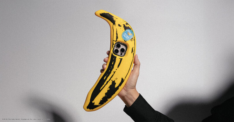 A hand holding a phone case shaped like a banana, with a camera lens cutout and a sticker on it.