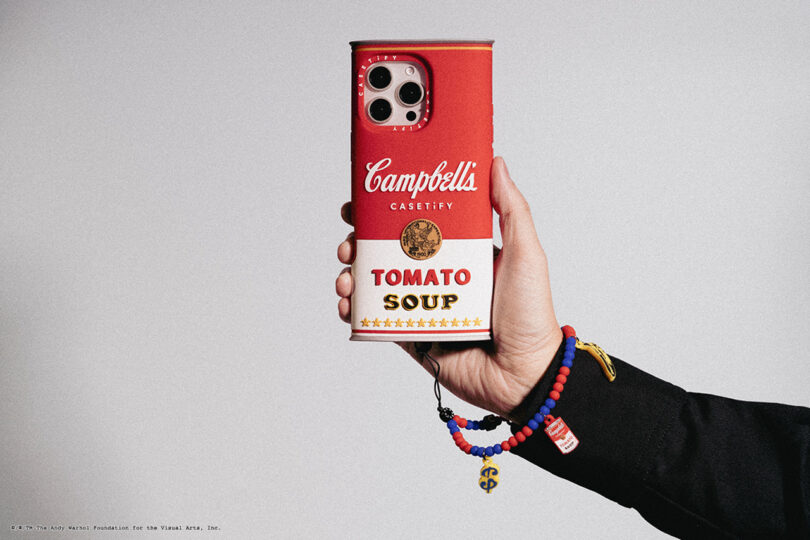 A hand holds a phone with a Campbell's Tomato Soup can design case. The person is wearing beaded bracelets.