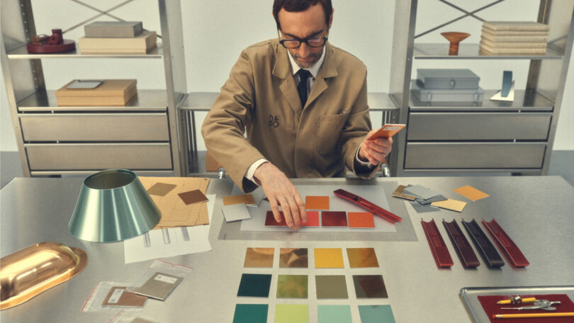 A man in a lab coat examines color swatches at a metal table, surrounded by materials and tools. Shelves with boxes and binders are in the background.