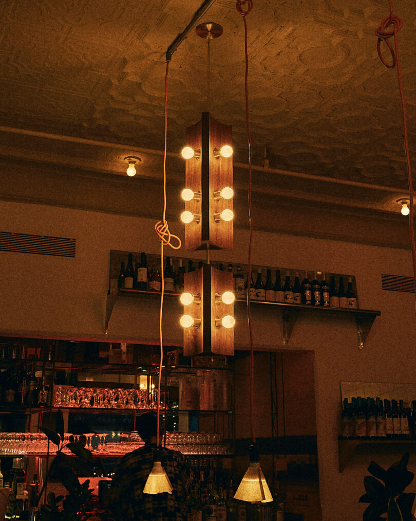 Dimly lit bar interior with hanging pendant lights, shelves filled with bottles, and a reflective bar counter. People are partially visible beneath the lights