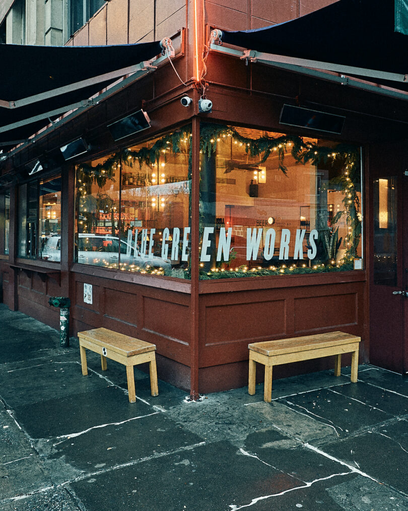 A corner cafe with large windows reading "LIKE GREEN WORKS," decorated with festive garlands, two wooden benches outside, and a wet sidewalk