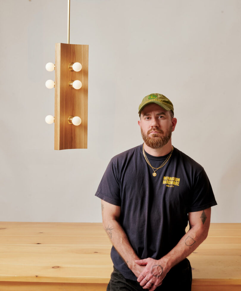 A man wearing a dark T-shirt and green cap stands in front of a wooden table. A modern wooden light fixture with round bulbs hangs above