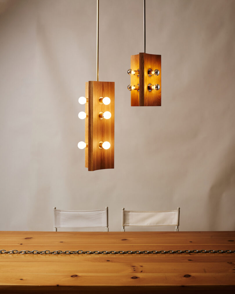 Two modern wooden pendant lights with exposed bulbs hang above a wooden table with a metal chain decor. Two white chairs are partially visible