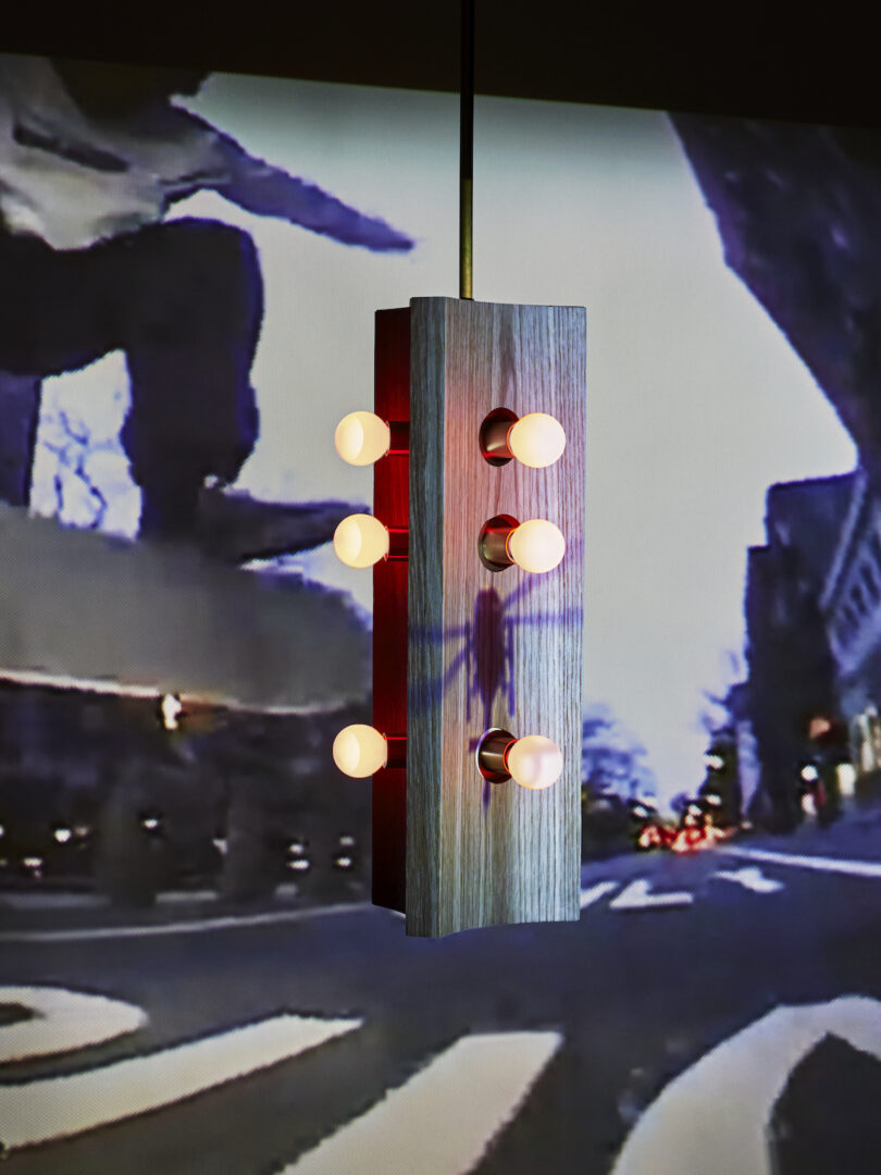 A hanging rectangular light fixture with multiple bulbs is in front of a projected image of a skateboarder on a city street